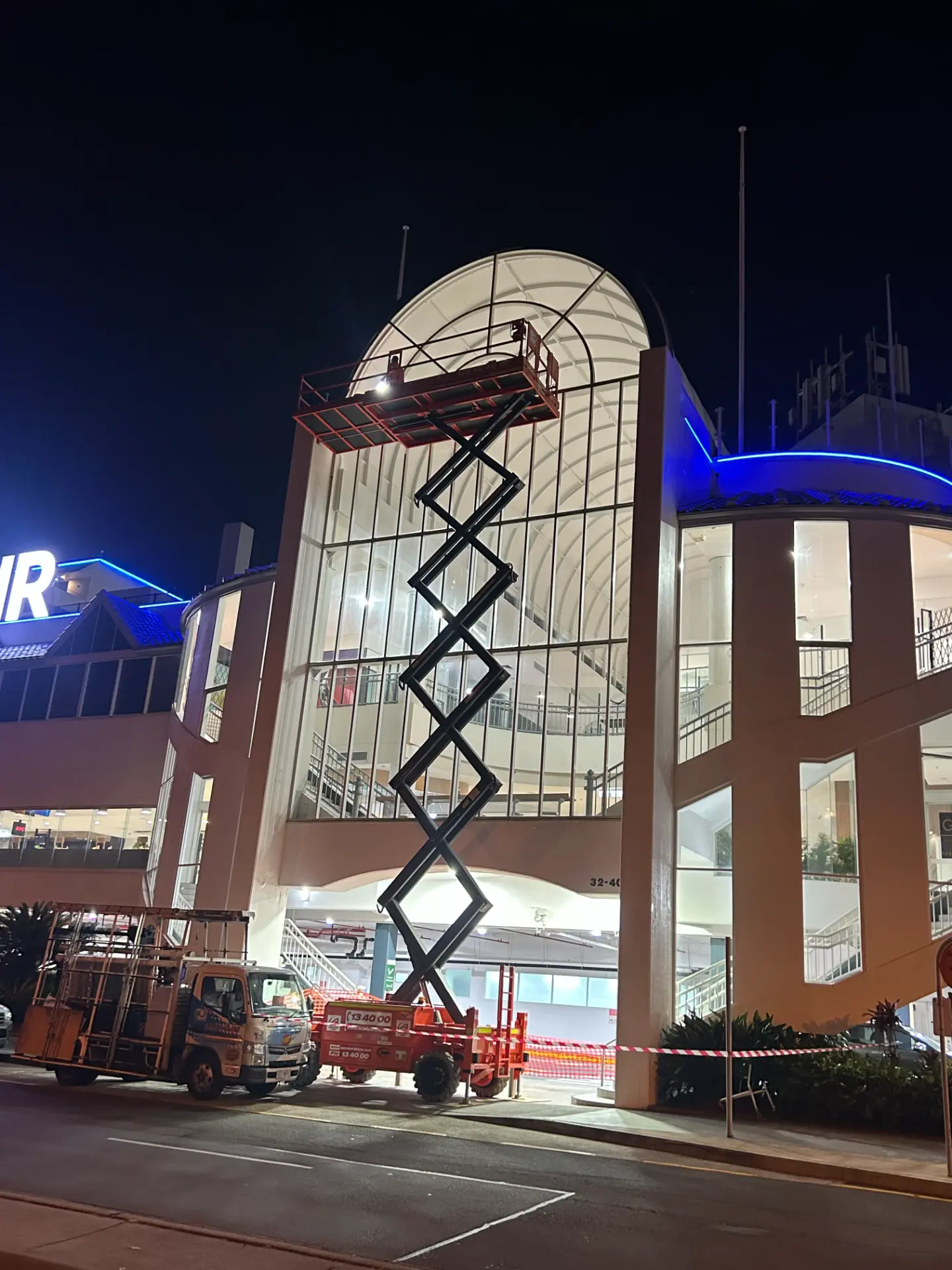 Scissor lift raised beside a commercial building's glass frontage at night